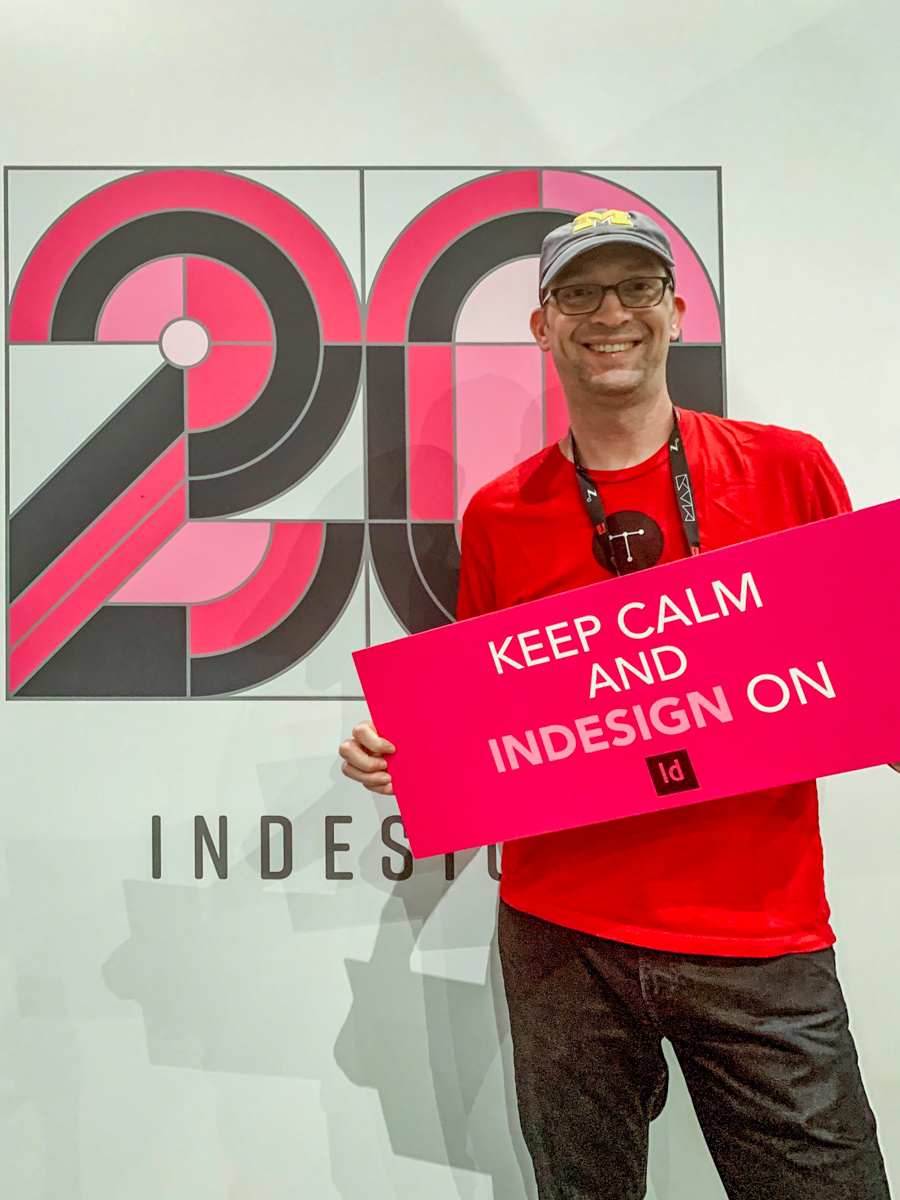 Caleb Clauset standing in front of a large banner displaying Adobe's official '20 years of InDesign' artwork. Caleb is wearing a t-shirt with the Typefi logo, and holding a sign that says 'Keep Calm and InDesign On'. He looks very happy!