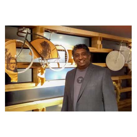 Chandi Perera stands in front of an artwork involving bicycles and fan-shaped pieces of cloth mounted on wooden racks.