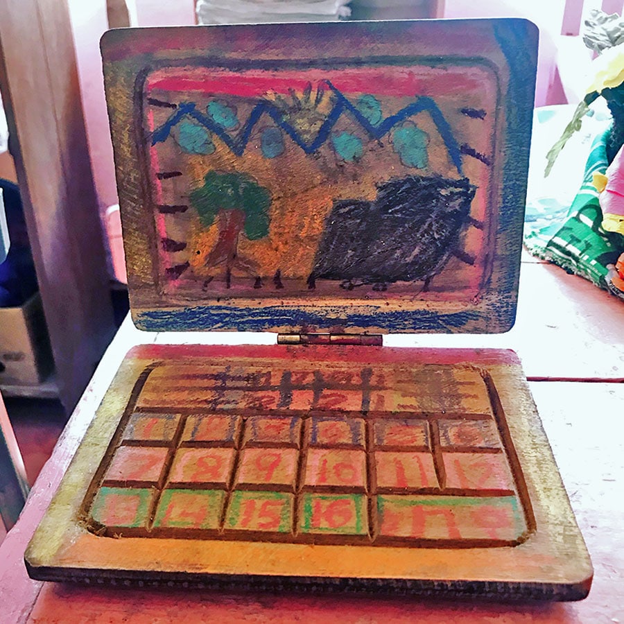 A toy laptop computer handcrafted from wood. The screen and keyboard are joined with a door hinge. A child has drawn a bright scene of trees and mountains on the screen section, and numbers on the keyboard section.