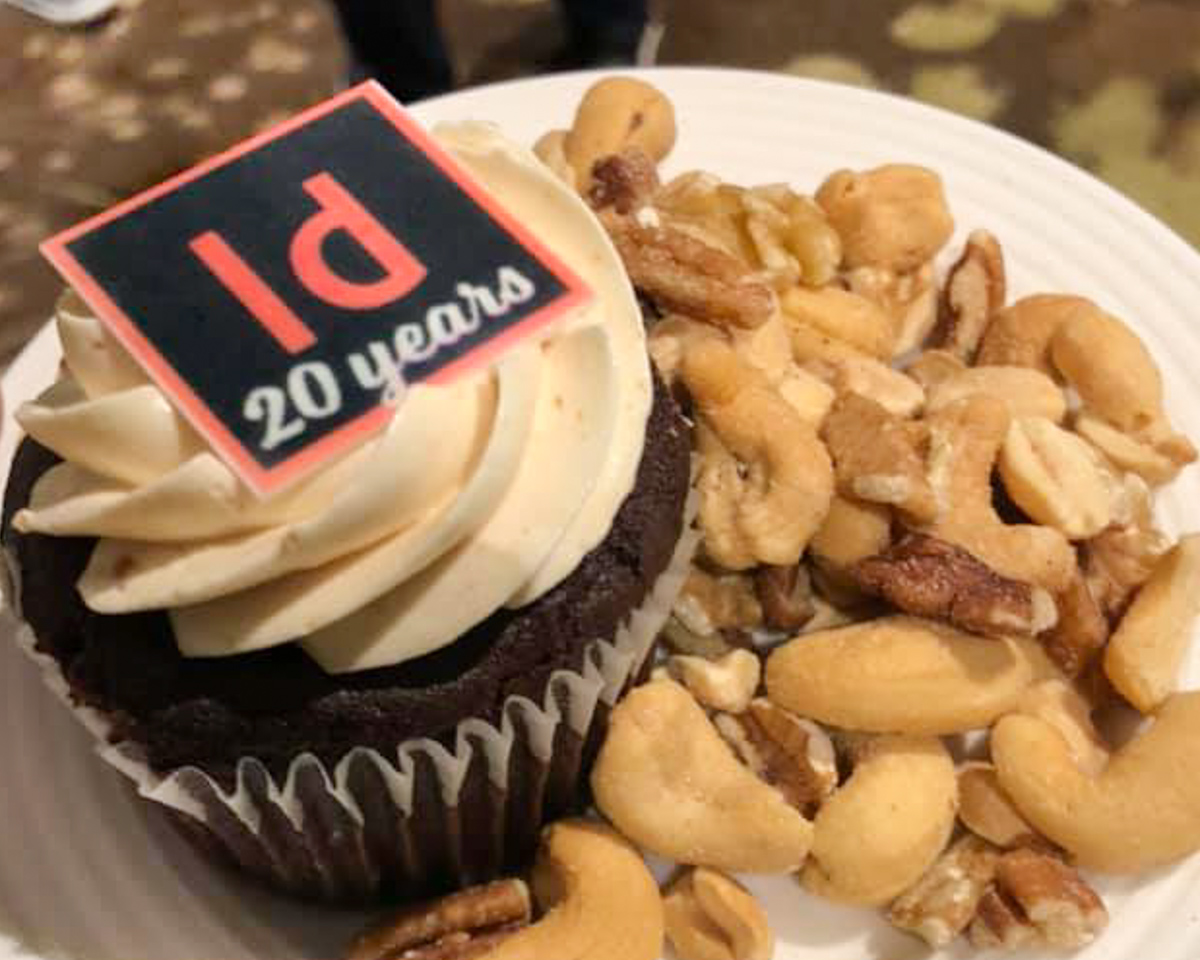 A delicious-looking cupcake with white icing and a black square decoration on top in the shape of the InDesign logo. The words '20 years' are written on the bottom of the logo. There is a pile of mixed nuts on the plate next to the cupcake.