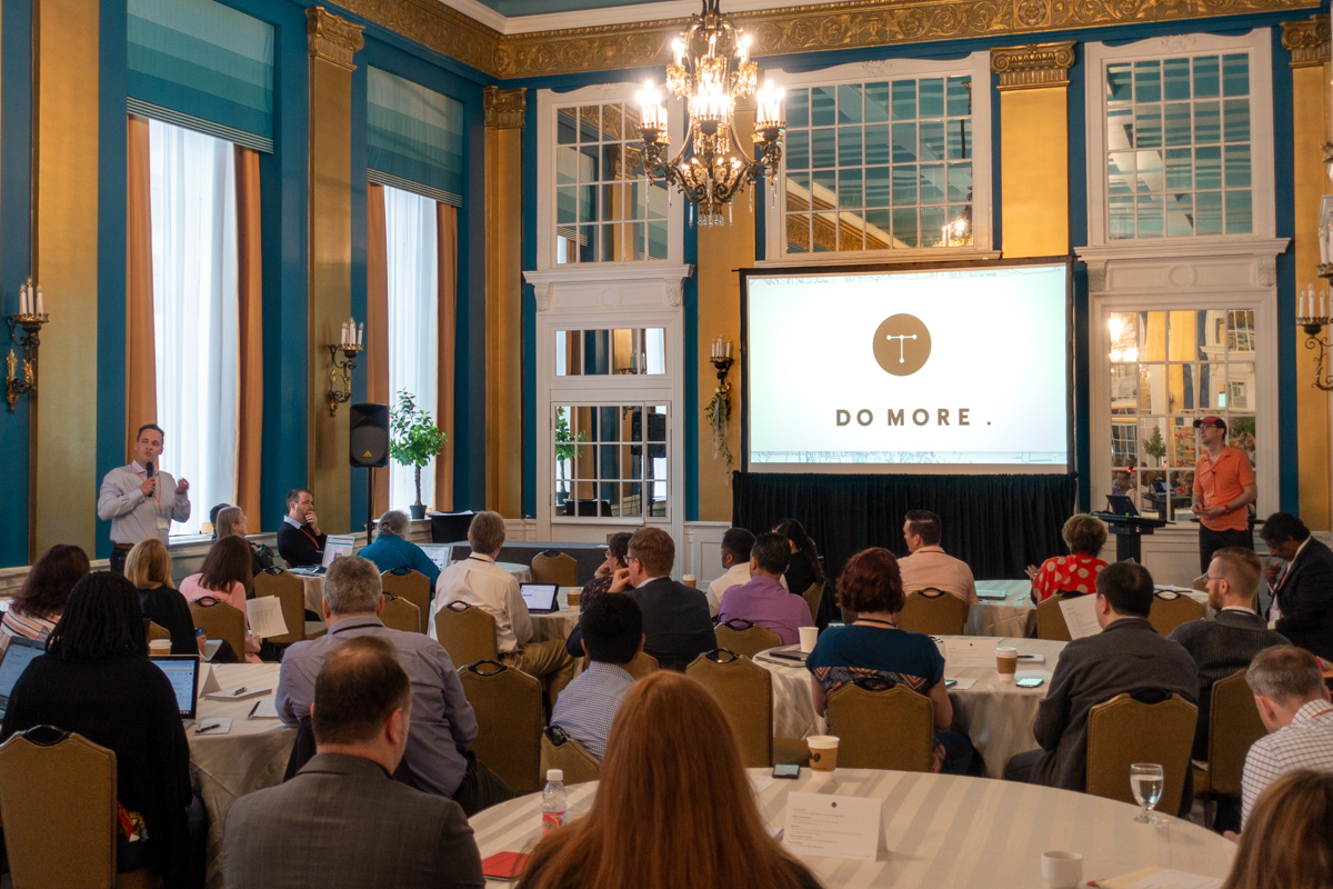 Guy van der Kolk and Caleb Clauset address the audience in the Versailles Room at Lord Baltimore Hotel during the Typefi User Conference. There is a large screen displaying the Typefi logo and the tagline 'DO MORE'.