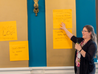 Christina Hampton gestures to two large post-it notes on the wall. The first one says 'Dashboard: Support tracking, ETA for deliverables, workflow management outside Typefi'. The second one says 'Tables keep options, subheaders, rows, table with one line'. There is a little sad face after the 'table with one line' comment!