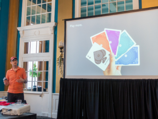 Caleb Clauset holds a deck of playing cards. A slide showing a hand holding up five cards (an ace and four different-coloured card backs) is displayed on the screen.