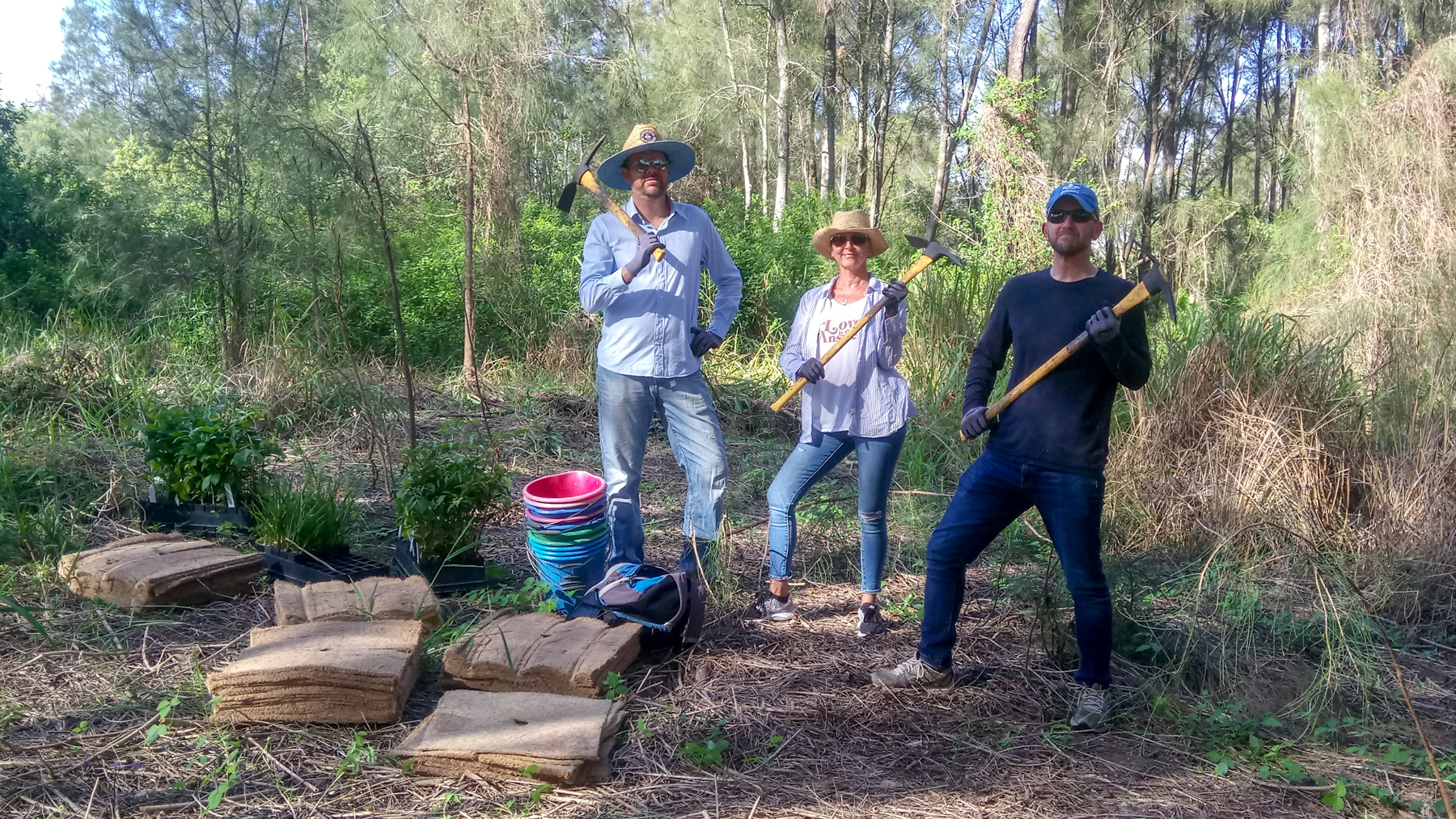 Ben, Sue and Michael wield mattocks in a cleared patch of bushland. There are piles of buckets, weed mats and seedlings on the ground next to them.