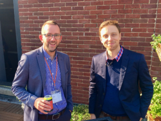 Michael Cousins and Tim Siers stand in front of a brick wall and greenery.
