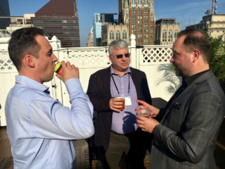 Guy van der Kolk, Robin Dunford and Stephen Laverick chat over drinks in the rooftop bar with other Baltimore buildings in the background. Guy is drinking from a soft drink can, and Robin is looking suave in sunglasses while Stephen makes a point.