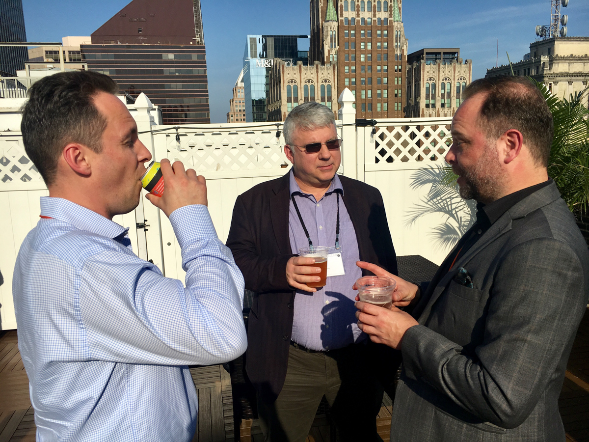 Guy van der Kolk, Robin Dunford and Stephen Laverick chat over drinks in the rooftop bar with other Baltimore buildings in the background. Guy is drinking from a soft drink can, and Robin is looking suave in sunglasses while Stephen makes a point.
