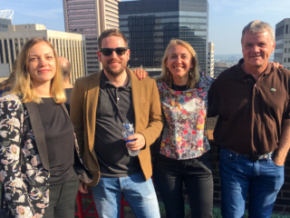 The group smiles at the camera near the wall of the sunny rooftop bar, with Baltimore visible in the background.