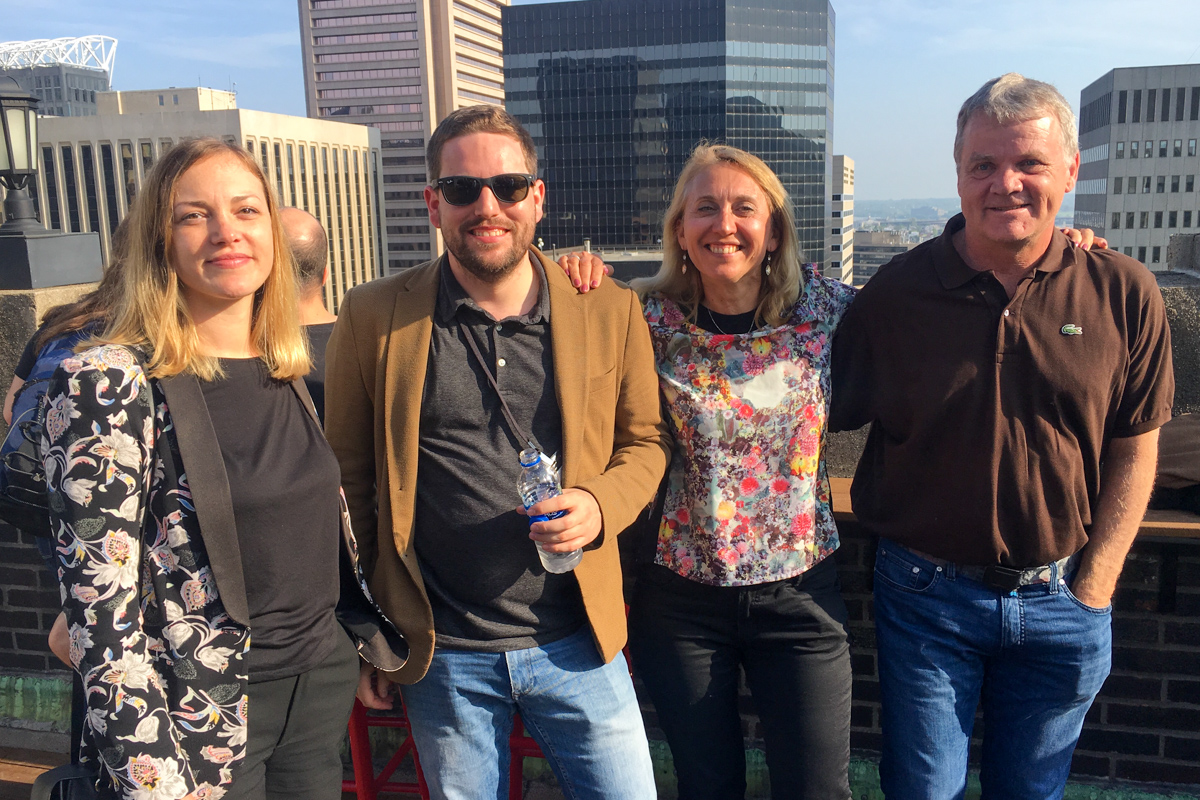 The group smiles at the camera near the wall of the sunny rooftop bar, with Baltimore visible in the background.