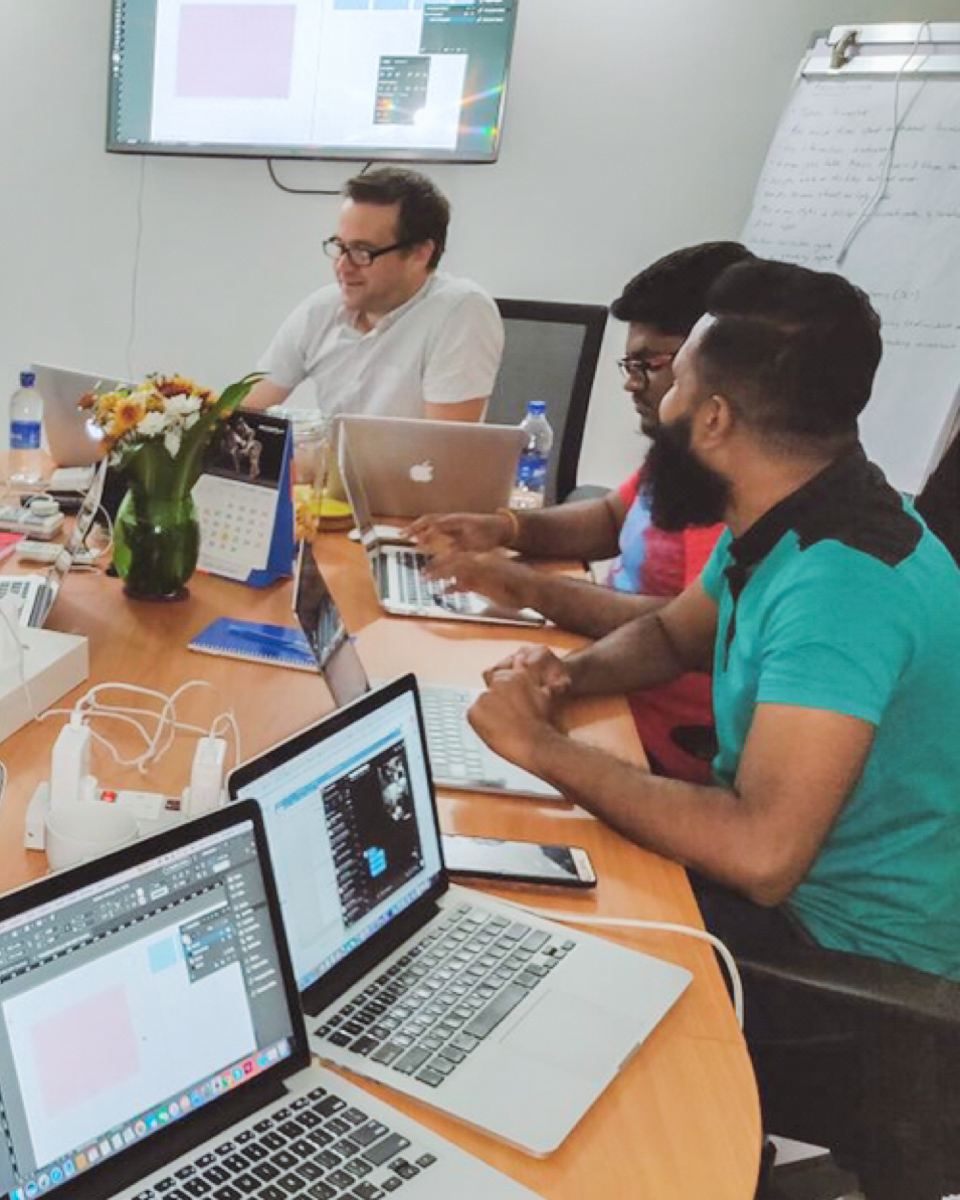 A training session in Typefi's Colombo office. Ken Jones is seated at the head of the table and has an InDesign layout projected on the screen. Dumesh Gajamange and Dilum Samarajeewa are seated at their computers, listening intently to Ken.
