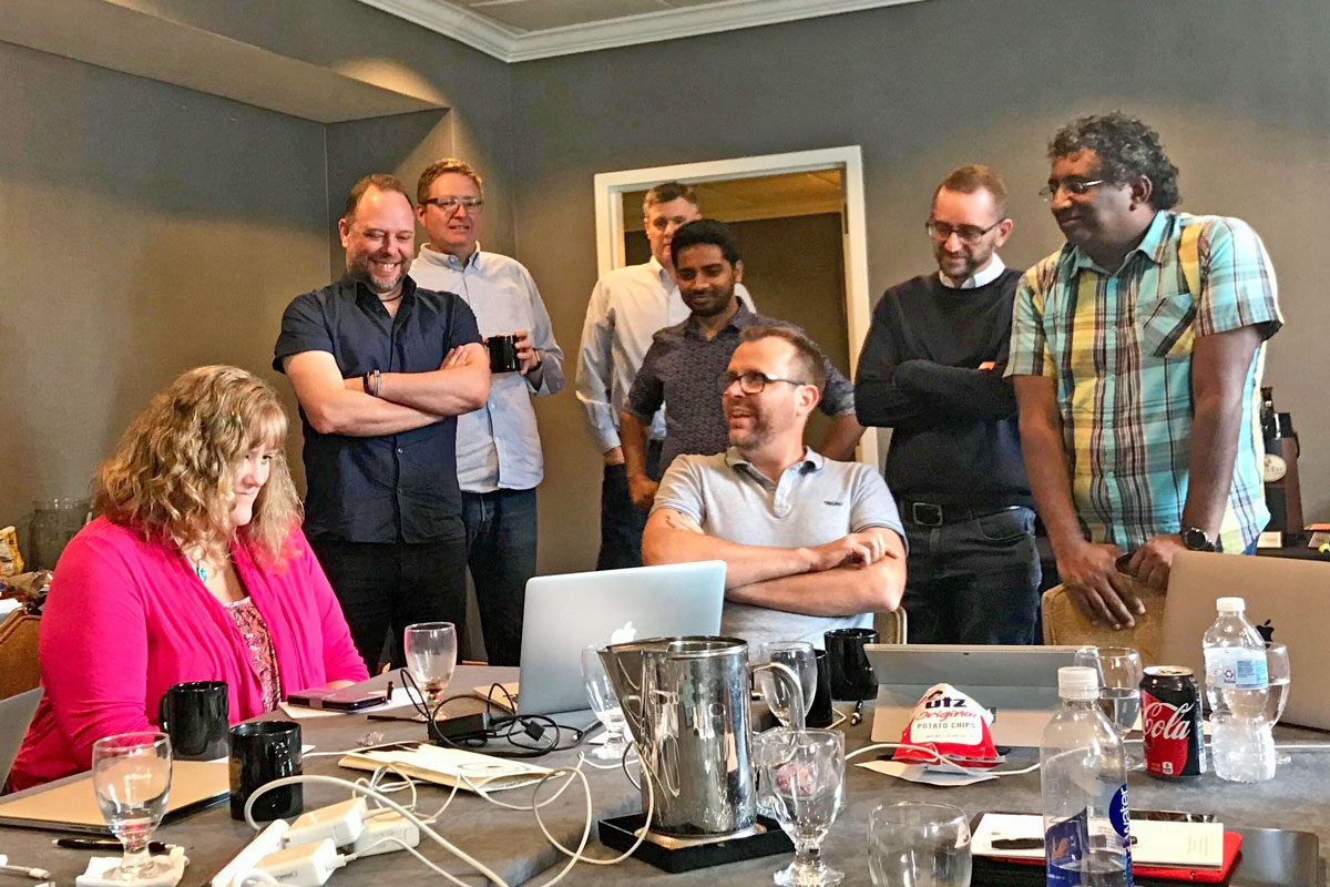 Eight members of the Typefi team standing around one end of a board room table, watching something on a screen and laughing.