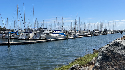 A marina with sailboats and yatchs docked. It is a bright, sunny day and there is water in the foreground.