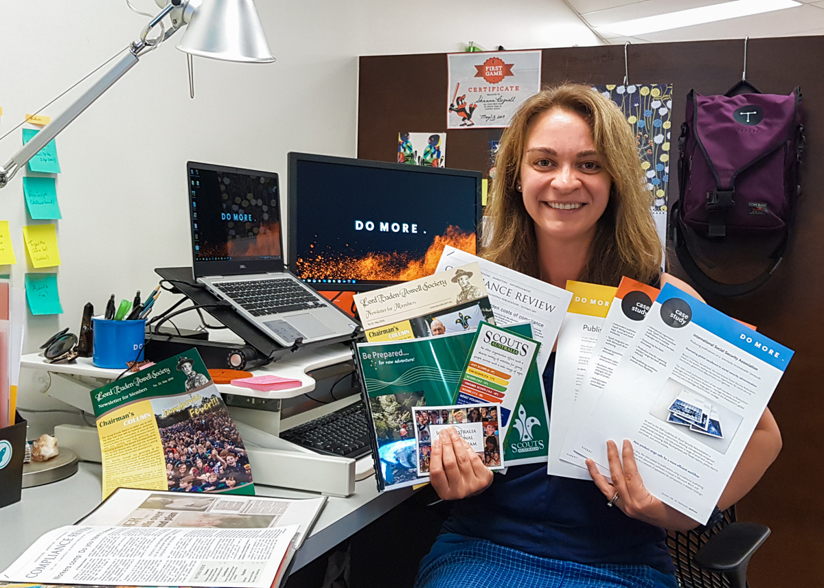 Shanna Bignell seated at her office desk holding a bunch of colourful brochures. The computer screens on her desk are showing the Typefi tagline 'DO MORE'.