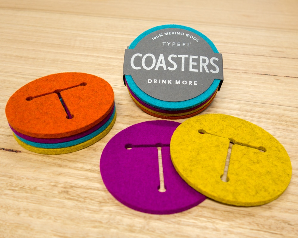A set of Typefi's 100% Merino wool felt coasters in orange, purple, yellow, and blue. Each coaster has a T-shaped cutout of the Typefi logo, and the label says 'DRINK MORE'.