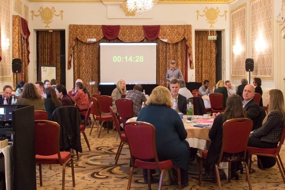 Groups of Conference attendees seated around tables discussing Typefi product features they'd like to have. There is a large clock counting down on the screen - there are 14 minutes and 28 seconds left.