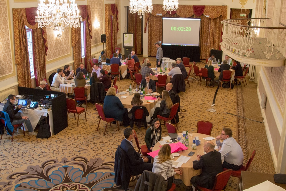 A shot of the Paganini Ballroom taken from an elevated position on one of the balconies. There are six groups of people in discussion around the tables, and the clock on the screen indicates that there are two minutes and 28 seconds left in the session.