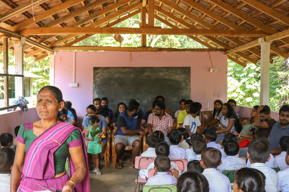 The Typefi team sits in front of assembled children at a Sri Lankan primary school while a teacher in brightly-coloured traditional dress looks on. The assembly area has an exposed wooden ceiling and just one wall with a chalkboard on it.