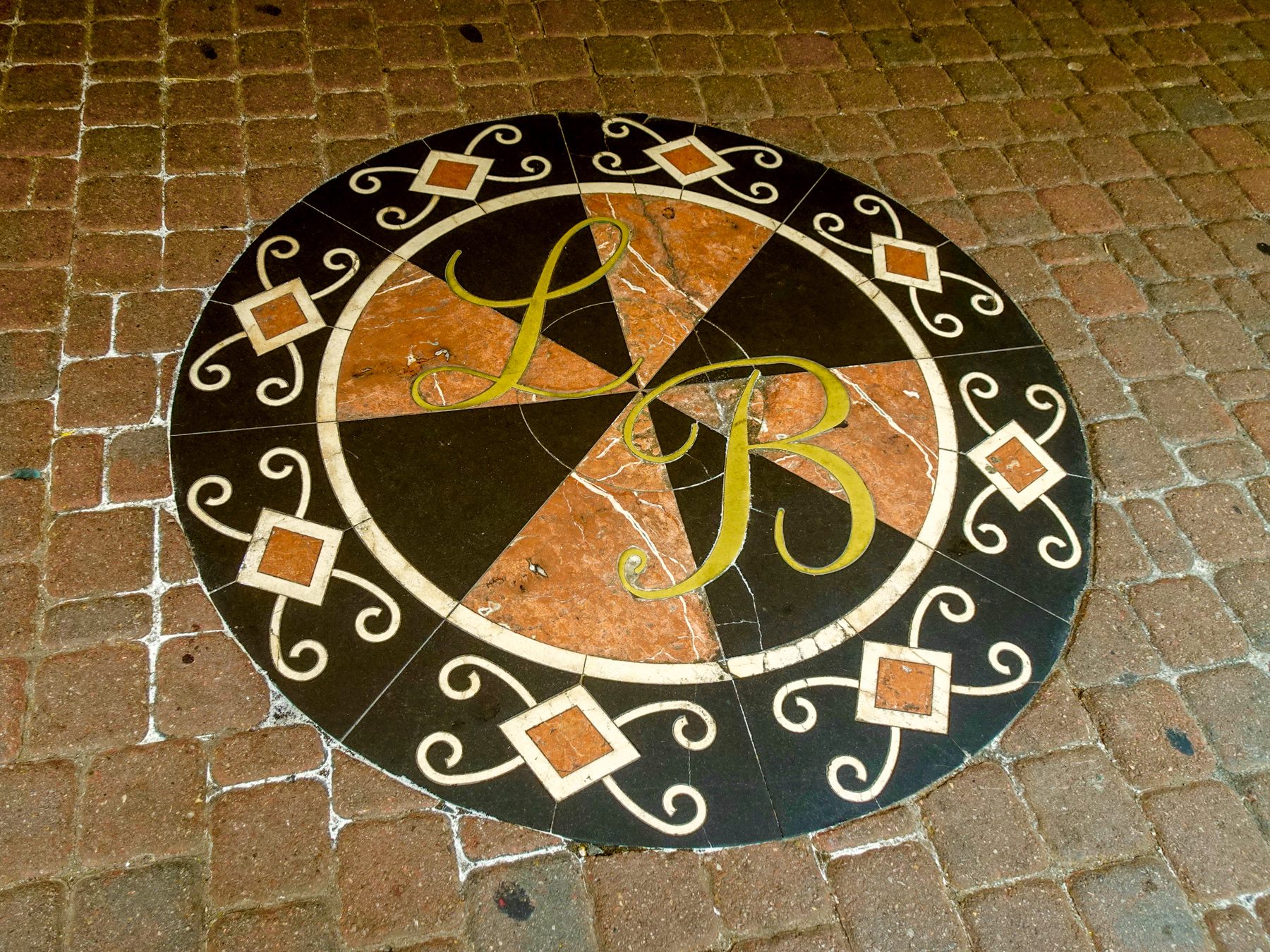 A circular mosaic set into the sidewalk, featuring the initials LB and decorative elements. The design is vaguely reminiscent of a dart board.